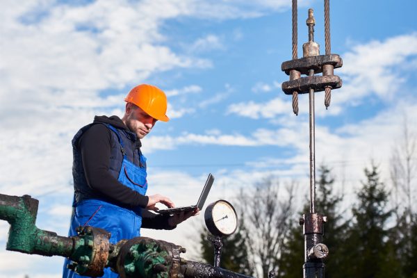 Engineer in work overalls and orange helmet checking oil pumping unit at oil field, using laptop. Male worker standing in front of oil well pump jack. Concept of petroleum industry, oil extraction.